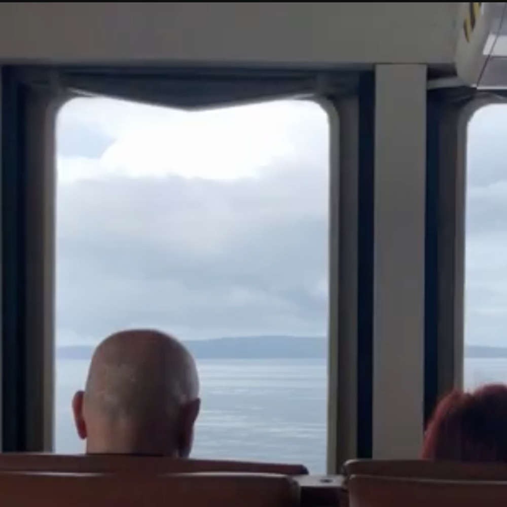 Una coppia seduta di spalle siede davanti ai finestrini di un traghetto. Snippet dell'intervista a Giacomo Deiana del Teatro dallarmadio per la rassegna Non è Teatro 2023.