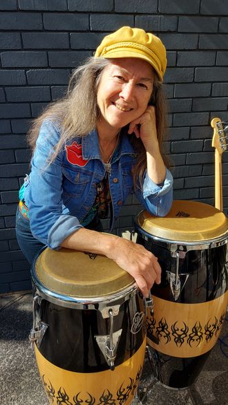 Kristine Best, looking cool, dressed casually in denim, wearing her trademark cap, with her congas (a conga and a kinto actually). She loves playing percussion.