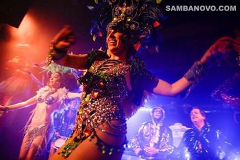 A dancer who is performing to live samba drumming under stage lights in a glittering sequined costume with a big smile
