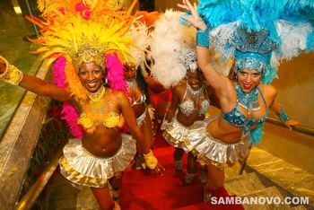 Four beautiful smiling Brazilian samba dancers in flamboyant yellow, pink, blue and white feathered costumes coming up stairs

