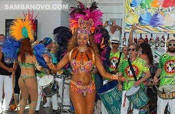 Two Brazilian dancers dancing samba as drummers play behind them The lead dancer is wearing an orange & pink Brazilian bikini
