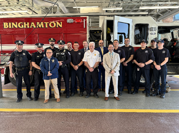 Image: Binghamton Police and Fire Departments, white men in black uniforms and hats, with Mayor Jared Kraham, a white man in a suit and tie, Philharmonic Executive Director Paul Cienniwa, a white man with dark glasses in a suit and tie, and Sales Director Brian Nayor, a white man in a tan suit and tie in front of a Binghamton Fire Department truck