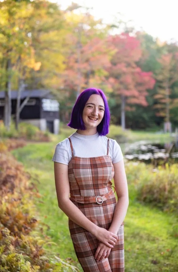 Alexis Drozdowski, a smiling white teenage girl with purple hair, standing in a landscape with trees and a small house