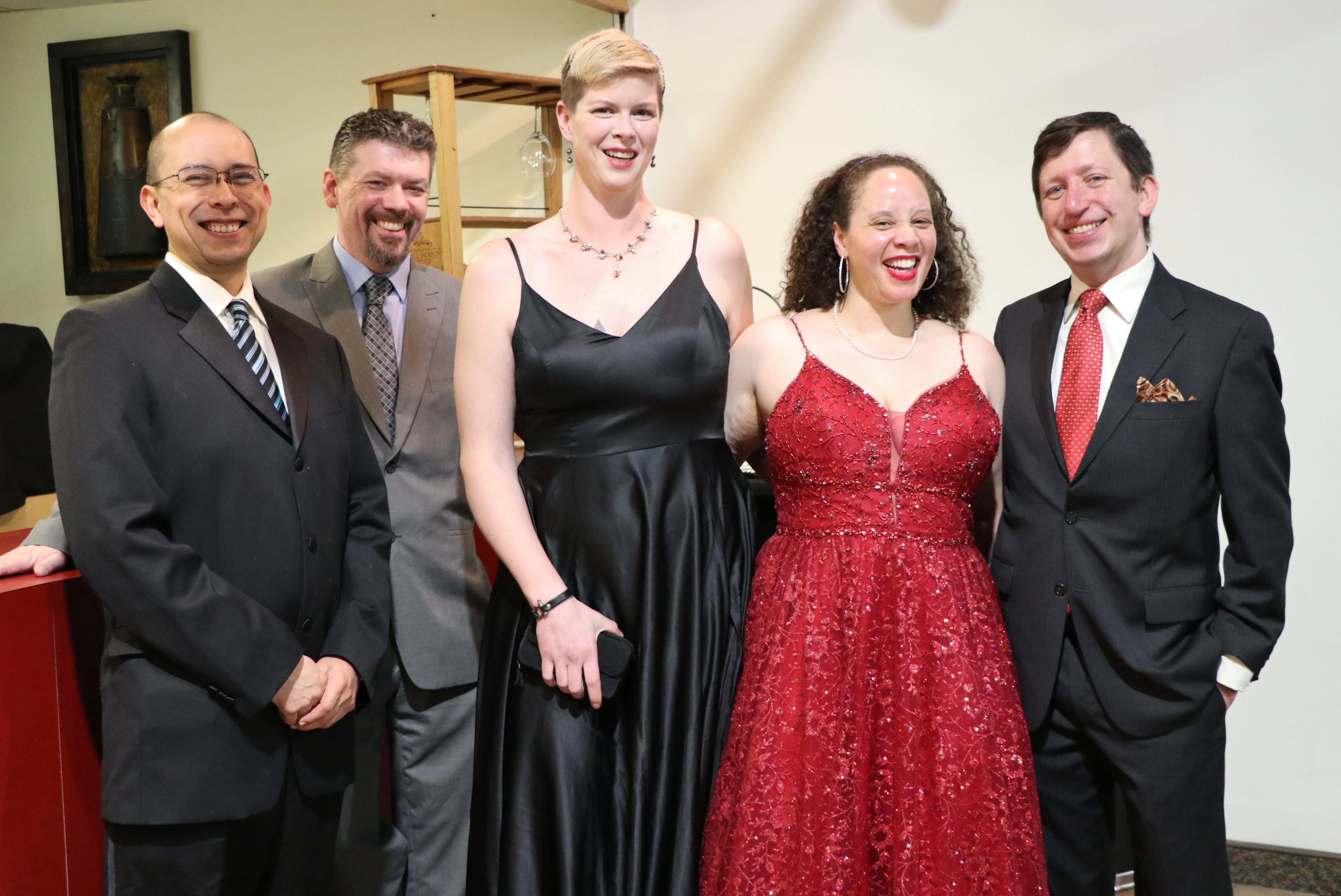 Carlos Perez, a bald Latino man with glasses in a suit and tie, Ajen Lewis, a white man with a beard in suit and tie, Cassie Lee, a tall white woman with short blonde hair in a black evening gown, Charlie Sobel, a biracial woman with long curly hair in a red evening gown, Joe Sobel, a smiling white man in a suit and tie