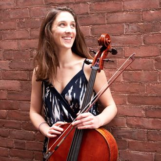 Image: Annie Jacobs-Perkins, a smiling white woman with long brown hair in black and white blouse holding a cello and bow in front of a brick wall