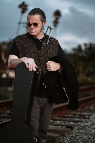 Photo of man in sunglasses by railroad tracks with harmonica and guitar case. 