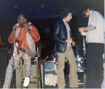 MUDDY, BOB DYLAN AND PAUL OSCHER MUDDY (BY MISTAKE) IS INTRODUCING BOB DYLAN AS "JOHN DYLAN". SOMEONE WHISPERED TO MUDDY "HIS NAME IS BOB, THATS BOB DYLAN" A FEW SECONDS LATER MUDDY CORRECTED THE INTRODUCTION AND THE CROWD WENT WILD. PAUL IS HANDING DYLAN A MARINE BAND TO USE WHILE HE SITS IN. (BOTTOM LINE NY 1975.) PHOTO: Willard Strandberg Jr..