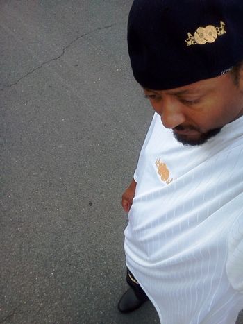 J.O.T. aka GRANDE GATO WEARING BLACK SOUL-FULL WEAR BASEBALL CAP WITH METALLIC GOLD LOGO, WHITE SHIRT WITH METALLIC GOLD LOGO, & BLACK KHAKI PANTS WITH METALLIC GOLD LOGO STANDING IN STREET ON BROKEN