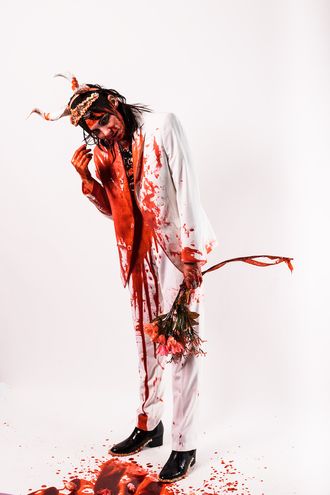 Medusa, trans nonbinary alt-pop musician and rapper; leaning, white suit, tuxedo, tiara, horns; bouquet of flowers; fake blood; prom scene, Carrie; photographer Justin Ruggiero