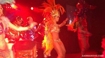 Samba dancers on stage in action dancing to live samba drummers who are behind them while an orange spotlight covers them all