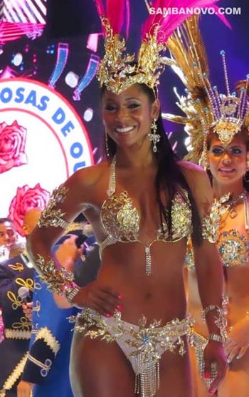 A dancer of samba on a stage who's wearing a vibrant, silver Brazilian bikini posing with her hand on her hip and a big smile