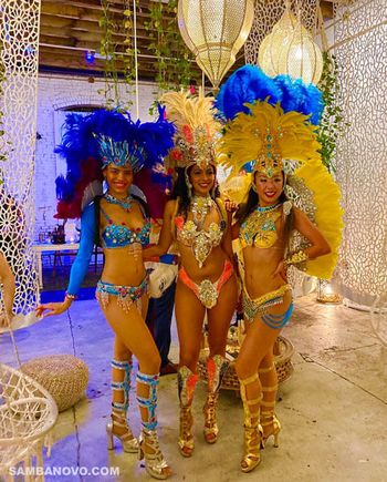 Trio of beautiful Brazilian dancers standing closely in a grand ballroom wearing blue, orange, and yellow Brazilian bikinis