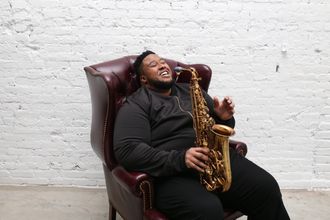 Musician & Model David Tv Barnes laughing while sitting in a red chair and holding a saxophone.