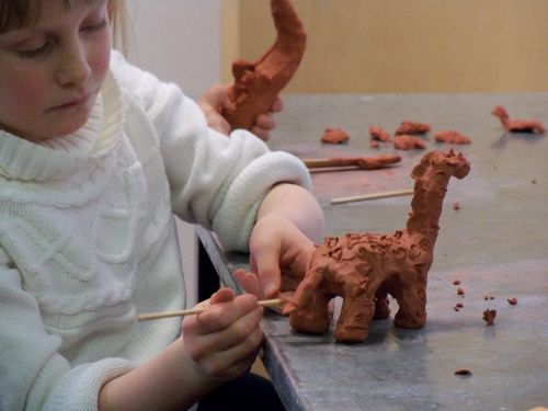 Young girl sculpts a giraffe from red clay in ARTistic Pursuits lesson links to homepage where parents can login to streamed courses. 