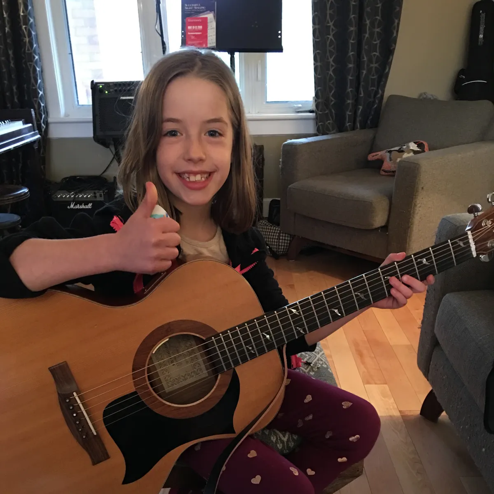 Young girl sitting and holding a guitar, while giving a thumbs up
