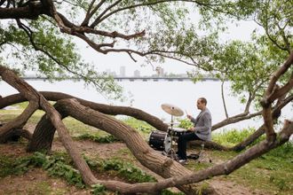 Dor Herskovits playing drums on the Charles river bank, surrounded by tree branches