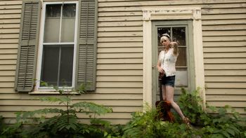 Woman standing outside house next to a guitar.
