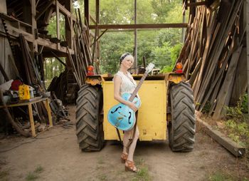 Woman standing outside in front of a tractor with a blue guitar.
