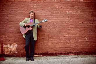 Motivational Humorist Greg Tamblyn and guitar by red brick wall