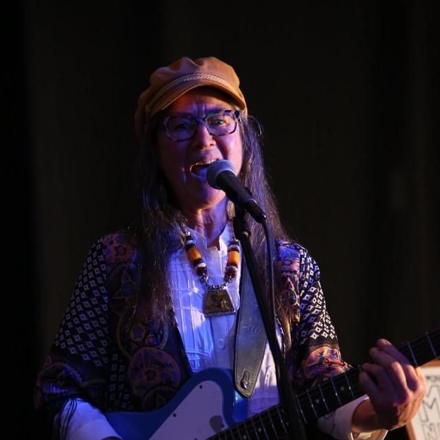 Kristine Best, singer songwriter musician and artist, is on stage playing her blue Telecaster and singing one of her original songs. She is wearing her trademark cap. 