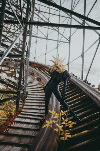 Photo of Medusa, non-binary pop artist, at abandoned theme park, wearing Venetian jester mask and clown ruffle collar. by Seekaxiom.