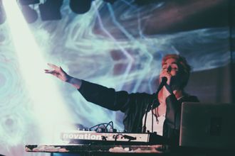 Medusa (musical artist) under a spotlight onstage at Mohawk Place, behind their synthesizers, performing Trail and reaching out their hand as they sing, in front of a projection of fractals.