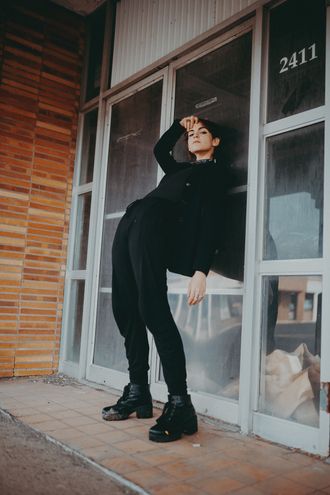 Medusa, non-binary pop musician and rapper, leaning against brick wall, hips forward, wearing black clothes and boots, showing jaw. Photo by Seekaxiom.