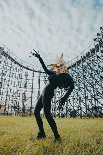 Photo of Medusa, pop musician, at abandoned theme park, in front of rollercoaster, leaning back,  wearing Venetian jester mask and clown ruffle collar. by Seekaxiom.
