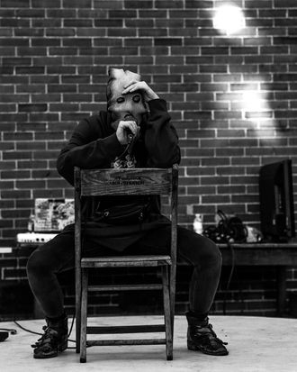 Photo of Medusa (musical artist) performing Danny Phantom onstage at a hip-hop festival. They sit backwards on a wooden chair in front of brick wall.