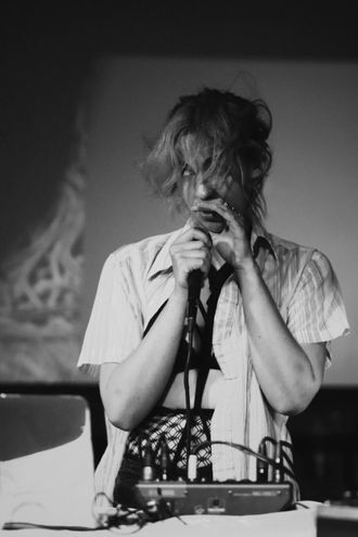 Medusa (musician) sings into a microphone, behind a mixer and laptop, looking to the side wearing fishnets and shibari under short sleeve button-down collared shirt.