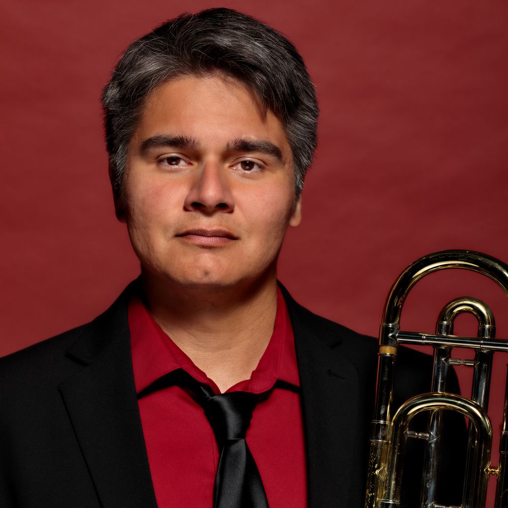 Headshot of Peter Tijerina holding a trombone. Black suit jacket, red shirt, black tie, and red background.