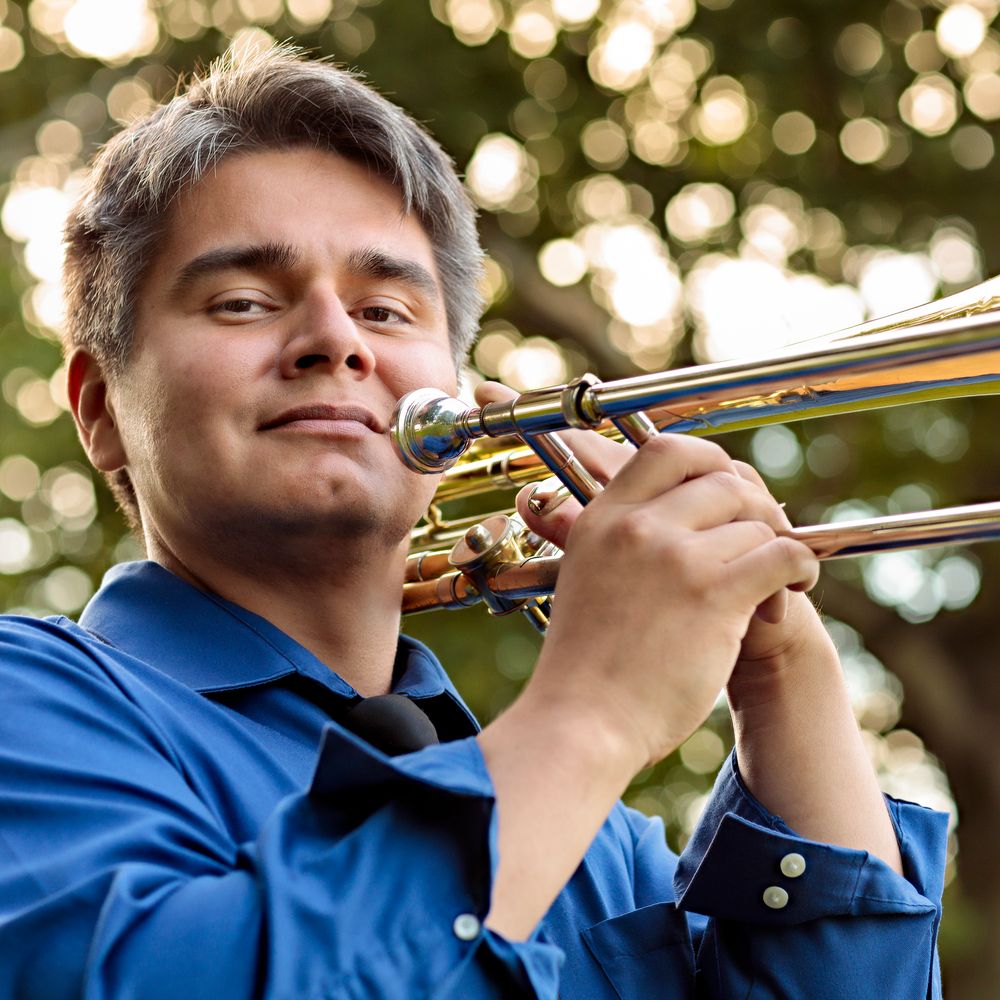 Photo of Peter Tijerina standing on a country road and holding a trombone. Wearing a blue shirt with a black tie and black slacks.