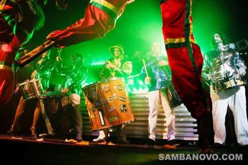 Samba drummers with large round drums strapped around them on stage playing live with green spotlights shining down on them
