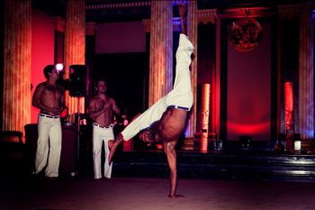 Three capoeira party performers in top form with shirts off, one of them standing on one hand while the others watch and clap
