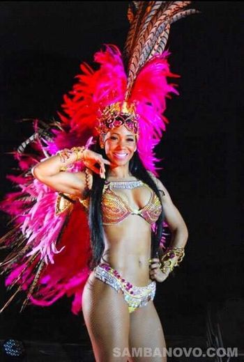 Hire samba dancers like this dancer who is wearing a red & magenta feathered costume while posing with one hand on her cheek
