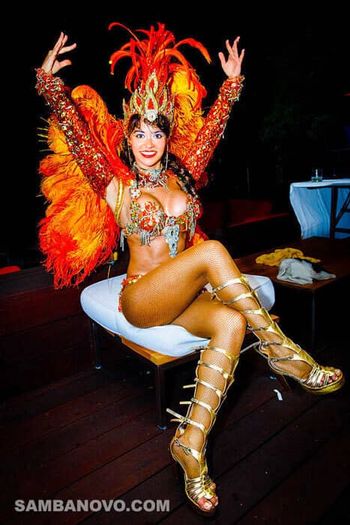 A gorgeous samba dancer sitting on a cushion with hands in the air wearing a shining orange costume with feathers on back
