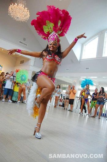 A Brazilian dancer in NYC who is samba dancing wearing a red costume while guests and the hosts watch and take pictures
