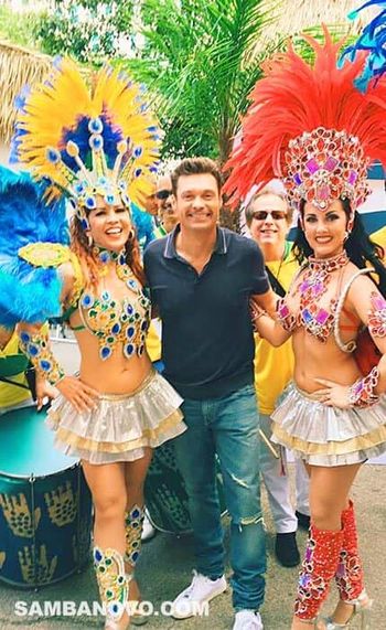 Ryan Seacrest standing in the middle of a samba group with two samba dancers at his side, all with big smiles in New York City
