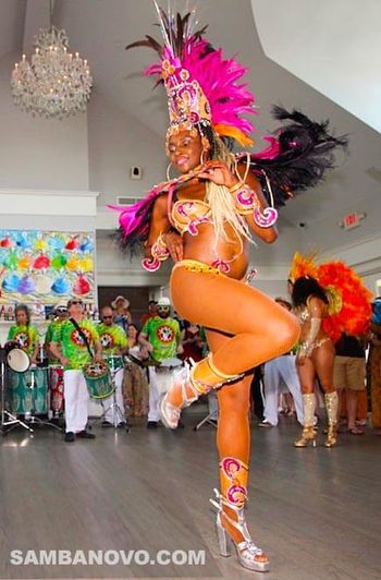 A beautiful dancer of samba dancing at a party wearing a pink and silver Brazilian bikini with live drummers playing behind her

