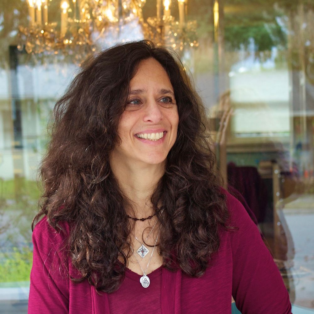 Long brown curly hair, smiling, looking towards the left, burgundy shirt and jacket, three necklaces. Standing in front of a window with mirrored images and Aunt Mariuccia's candelabrum.