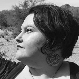 Light-skinned Mestiza with short dark hair looks intently to the side. An earring with a concentric circle design swings below the hairline. The backdrop is a mountain, cacti, and desert tree