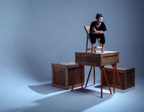 pale woman with dark hair and bangs seated on a chair on top of a box on top of an orange sawhorse. 