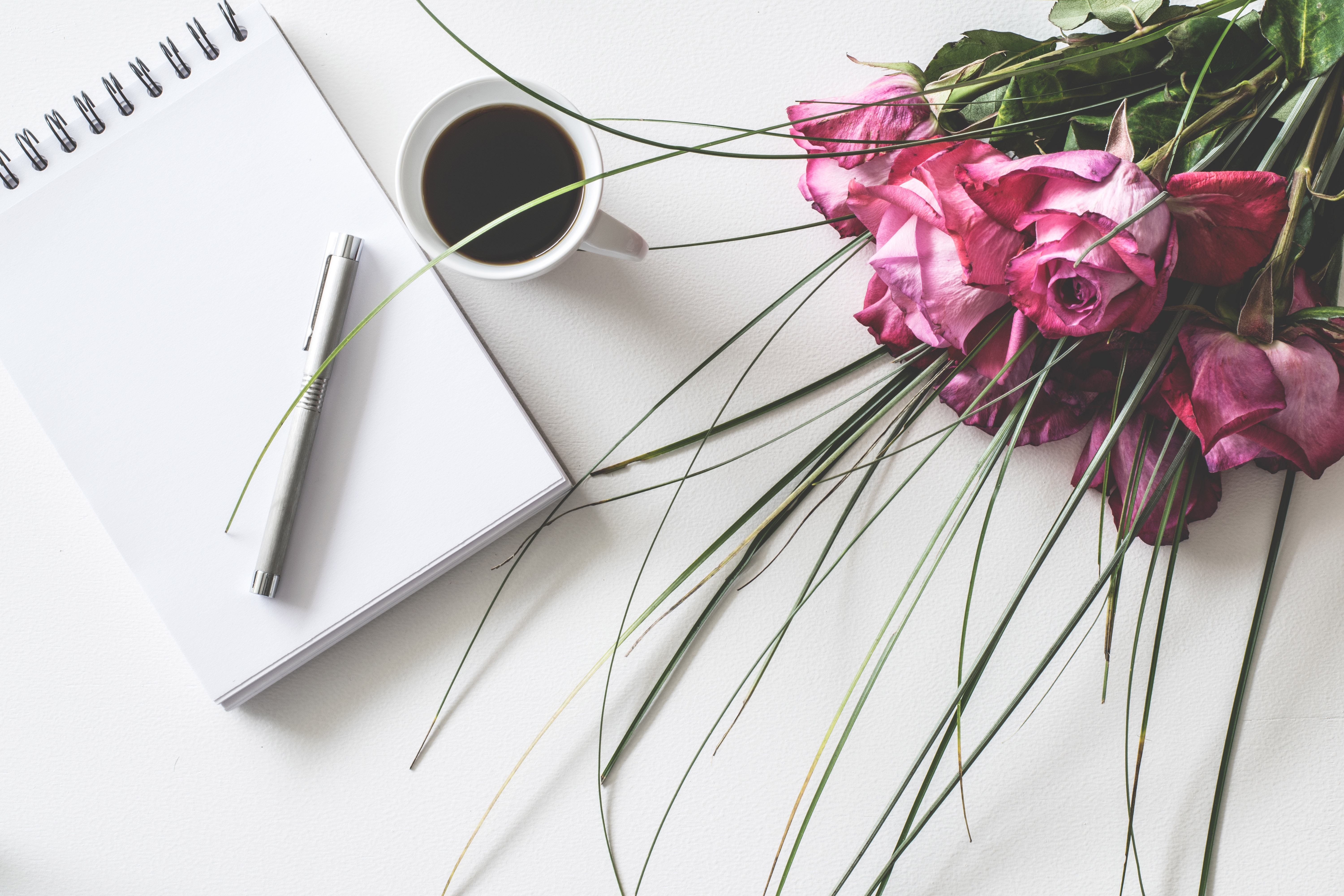 A beautiful bouquet of red roses sits beside a book, pen, and coffee cup on a white surface.