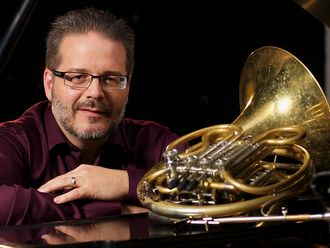 Image: Alex Shuhan, a white man with glasses sitting at piano on which a French horn is resting