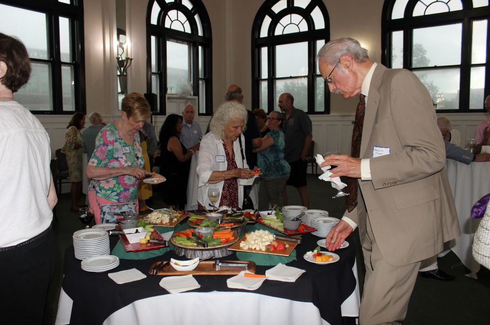 Two white women and a white man at a buffet table with fruits, vegetables, and cheeses. Other patrons are clustered behind them.