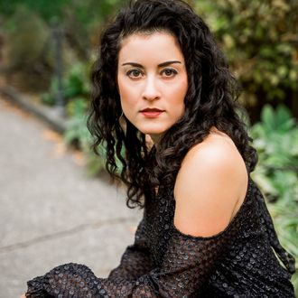 Image: Hilary Purrington, a white woman with long dark hair in a black blouse sitting on a sidewalk in front of a green bush