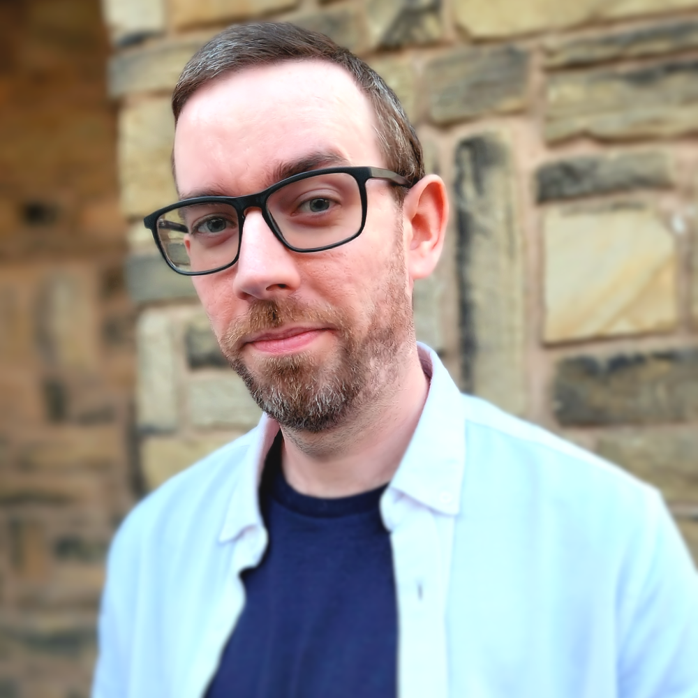 Photo of SEANIO in front of a blurry, aged sandstone brick wall; he is wearing an open white shirt with a navy blue top underneath and a pair of black rimmed glasses.