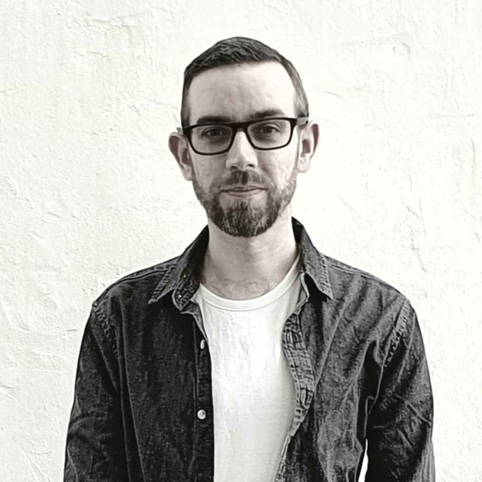 SEANIO main promo photo; image of SEANIO against a white textured background, wearing an open grey denim shirt and a white t-shirt underneath, as well as a pair of black rimmed glasses.
