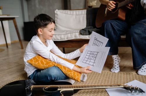 young boy smiling sitting on floor holding guitar tablature lesson with guitar laying on ground next to him