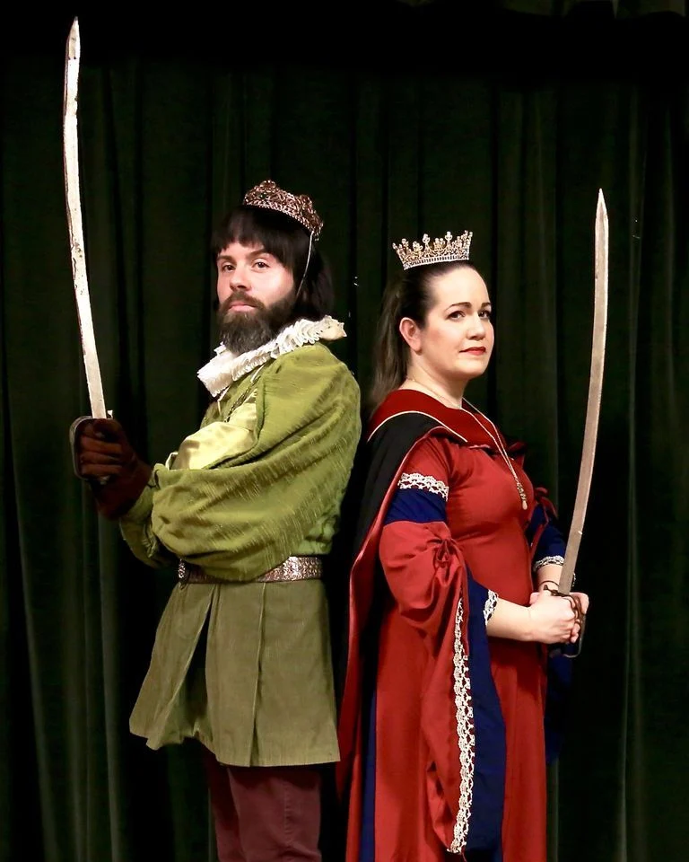 The picture below from Left to Right: Joseph Anthony Smith (Prince Hilarion) and Kara Vertucci (Princess Ida). Dark curtain background. Prince Hilarion is a man who has a dark brown bob and full and sculpted beard He wears a ruff, green tunic and red pants with matching red gauntlets and a bejeweled bronzish crown, and Princess Ida, a woman with long tied back brunette hair. She wears a gold necklace, red dress adorned with gold accents, a blue trim, and a black and red academic dress, and atop her head a tall gold crown. Both, crowned, stand back to back, regally, wielding swords pointed upright.